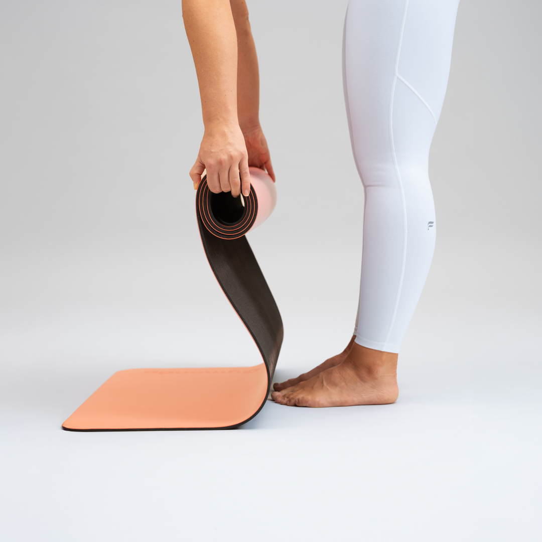 gebügte Person in weißen Yogaleggings rollt eine Gymondo-lachsfarbene Yogamatte auf, die teilweise noch auf dem Boden liegt