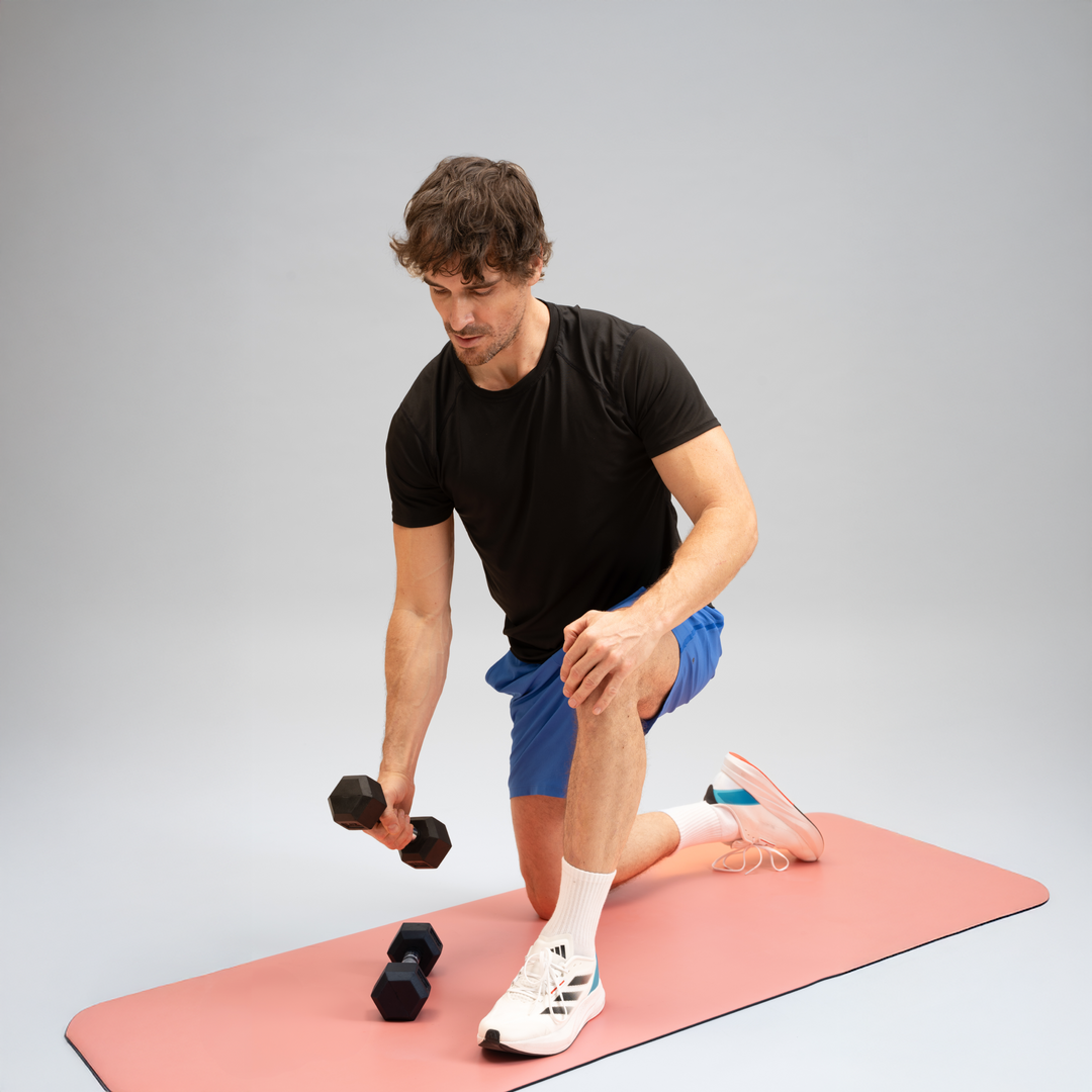 Männlicher Gymondo-Trainer in schwarzem T-Shirt mit Kurzhantel in der rechten Hand auf lachsfarbener Gymondo-Trainingsmatte